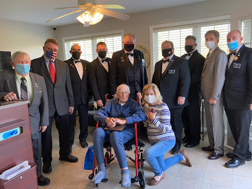 A formal gathering with an elderly individual in a wheelchair, surrounded by men in suits and women, all wearing masks.