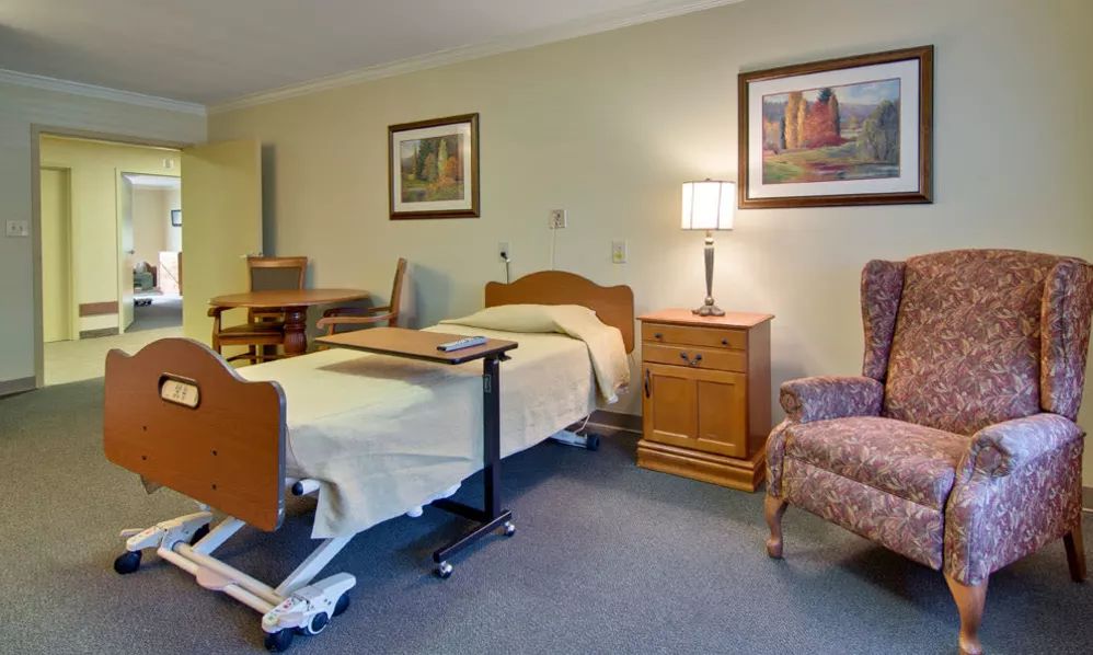 A hospital room with a bed, side table, chair, and lamp.