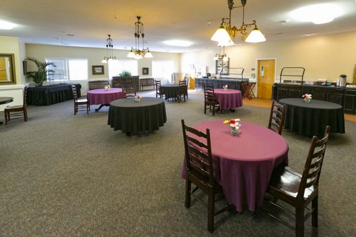 A spacious dining area with round tables and wooden chairs, featuring chandeliers for lighting.