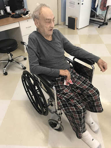 An elderly individual sitting in a wheelchair, dressed comfortably in plaid pants.