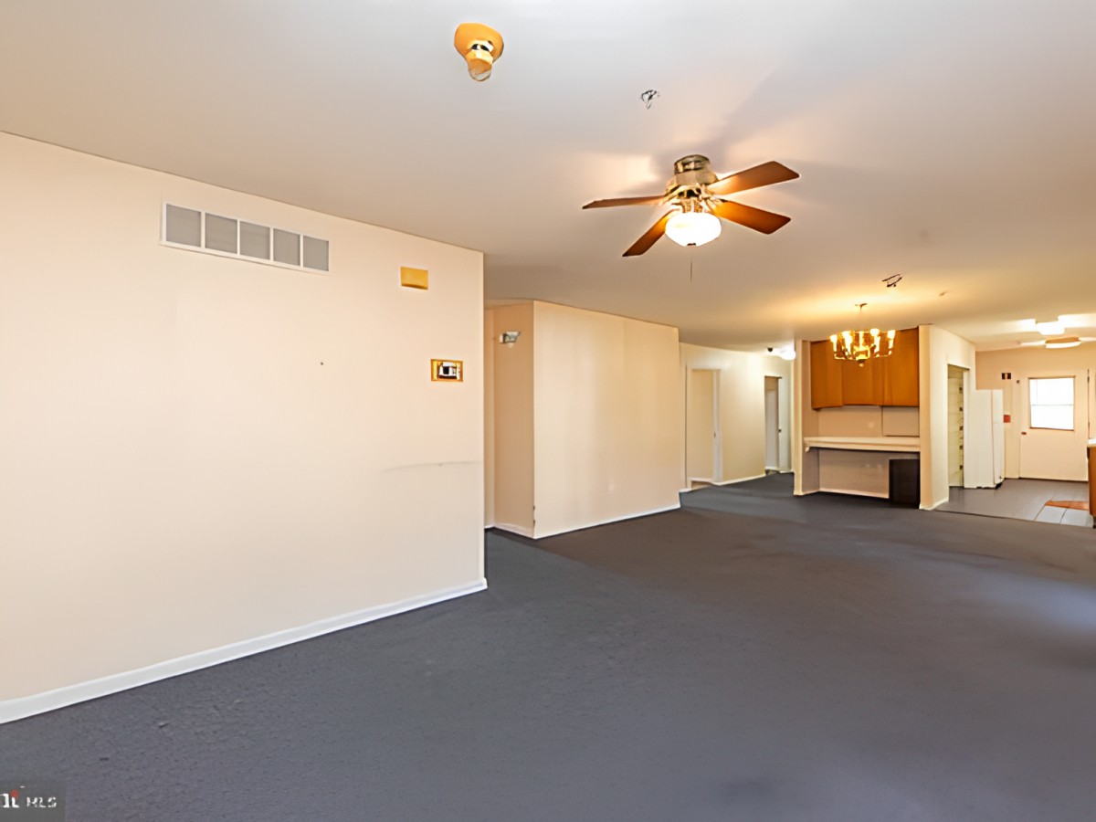An empty living room with blue carpet and a ceiling fan, leading to a kitchen area.