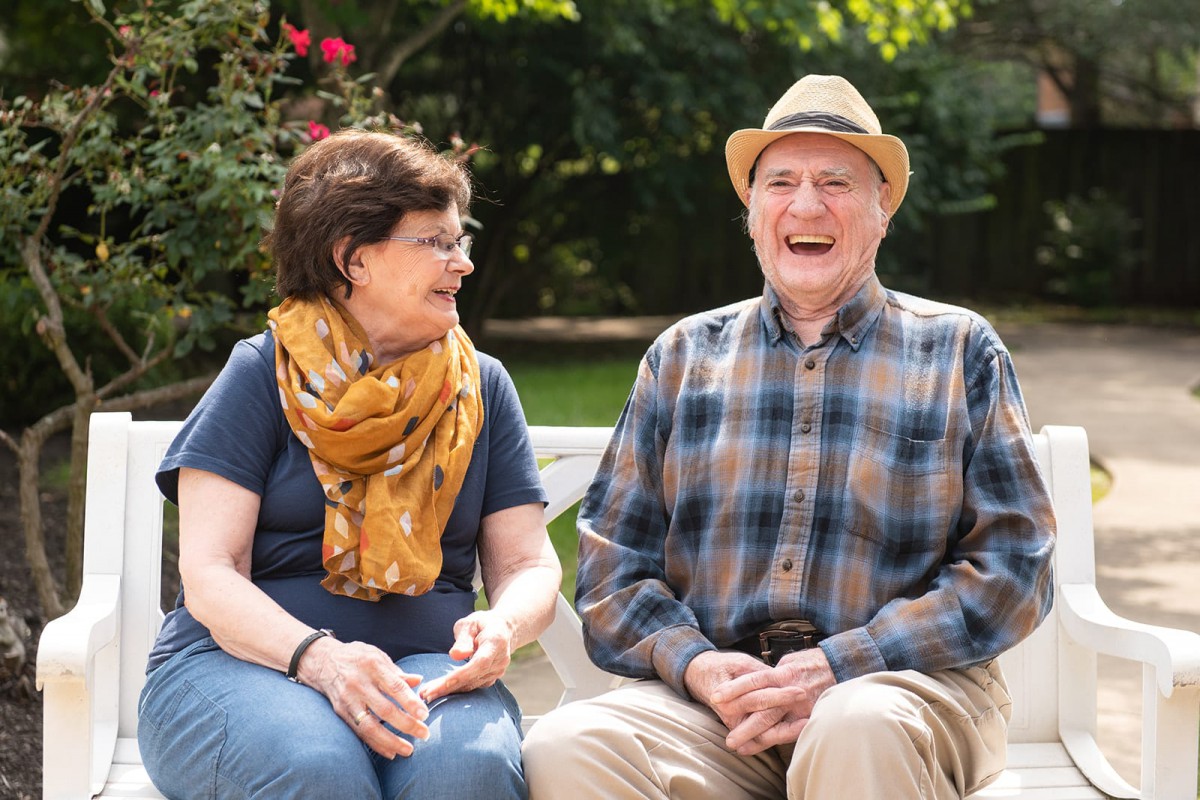 An elderly couple enjoying a sunny day in the garden, sharing laughter while sitting on a bench.