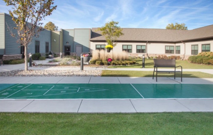 An outdoor shuffleboard court in front of a modern building, surrounded by well-maintained landscaping on a sunny day.