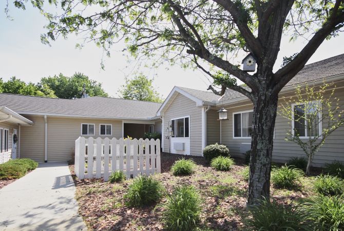 A single-story house with a front yard featuring landscaping and a white picket fence under a large tree.