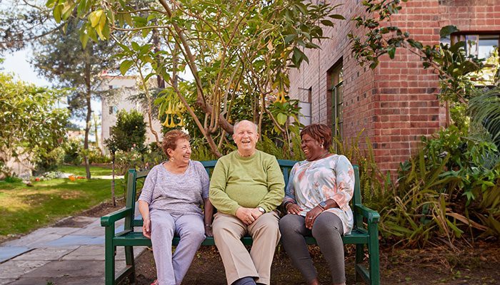 Three elderly people sitting on a bench in a garden, enjoying each other's company.