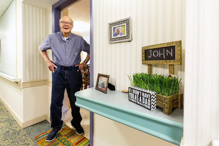 A smiling older man stands in a doorway next to a personalized shelf with decorative items.