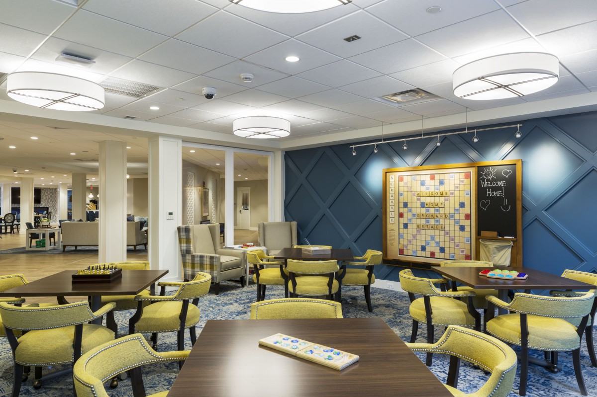 A modern game room featuring tables and chairs arranged for playing board games, with a large Scrabble board on the wall.