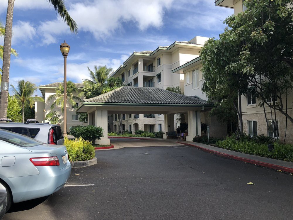 The entrance of a hotel with palm trees and parked cars under a partly cloudy sky.