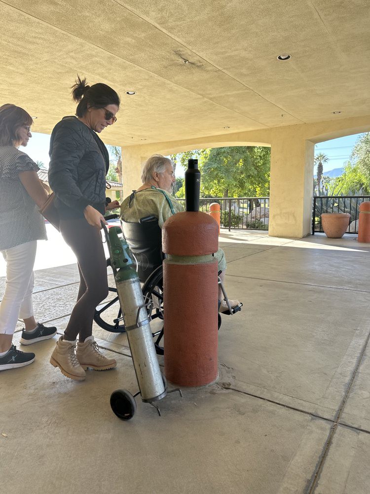 A woman in a wheelchair with an oxygen tank being assisted outside by two caregivers.