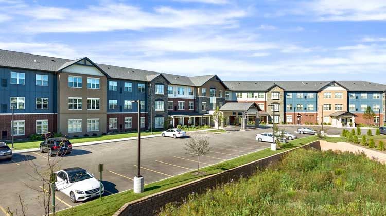 A modern three-story residential building with a spacious parking lot under a partly cloudy sky.