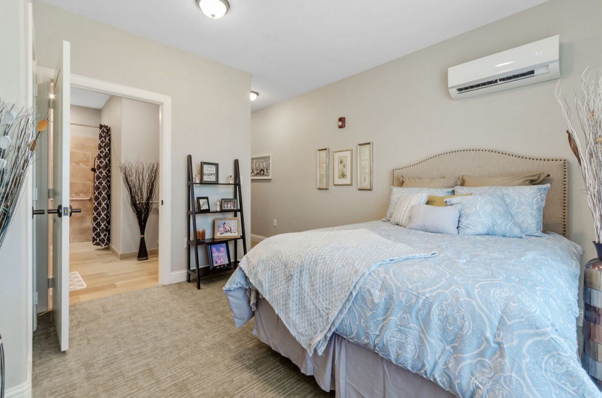 A cozy bedroom with a neatly made bed, shelves with decor items, and an air conditioning unit on the wall.