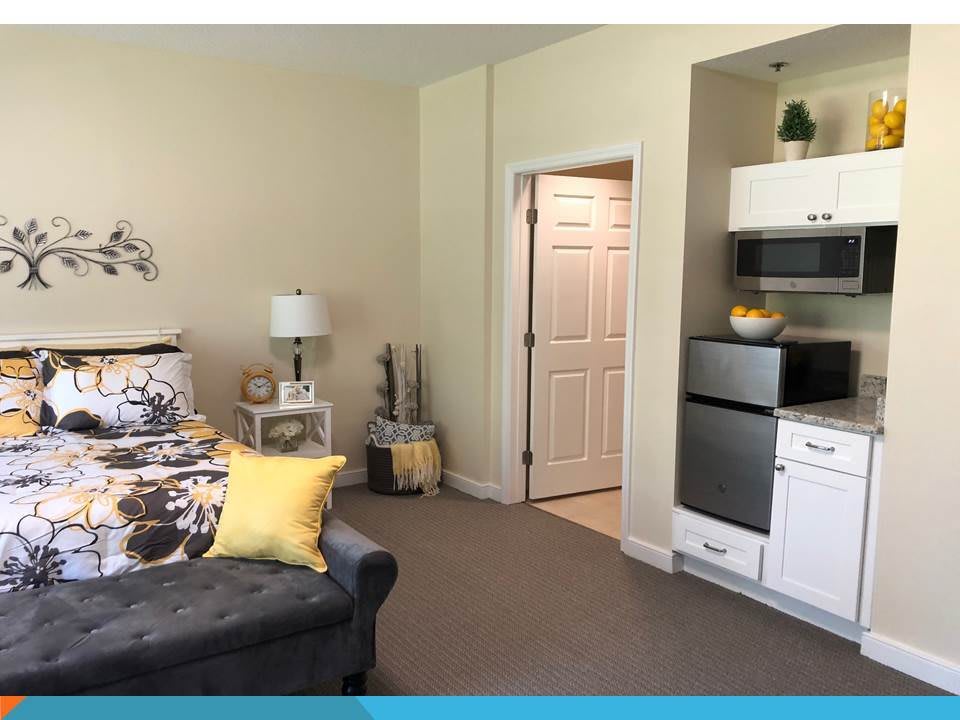 A cozy room featuring a sofa with floral and yellow pillows, a lamp on a side table, and a kitchenette in the corner.