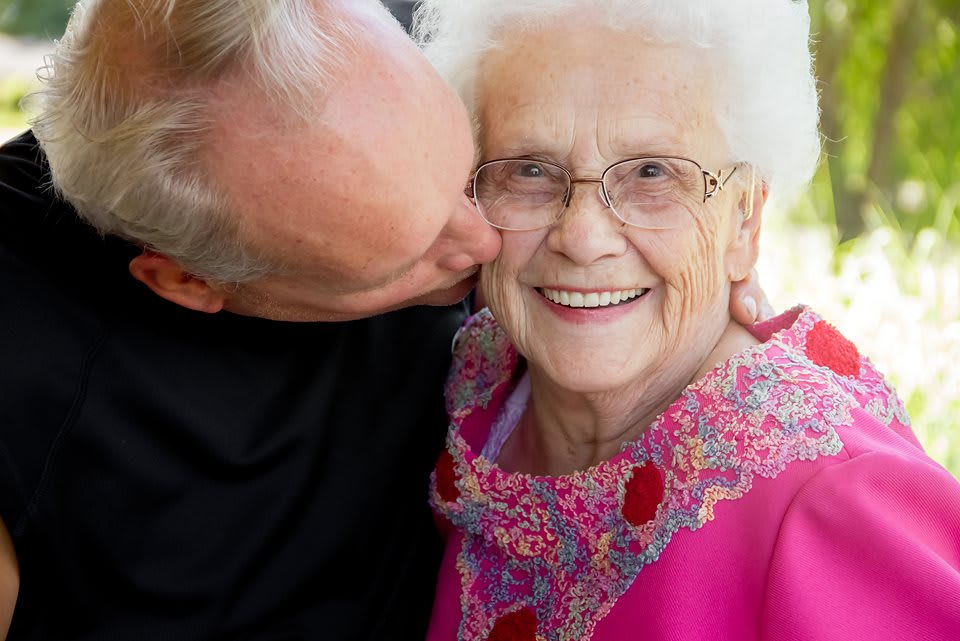 A man affectionately kissing an elderly woman on the cheek, both smiling warmly.