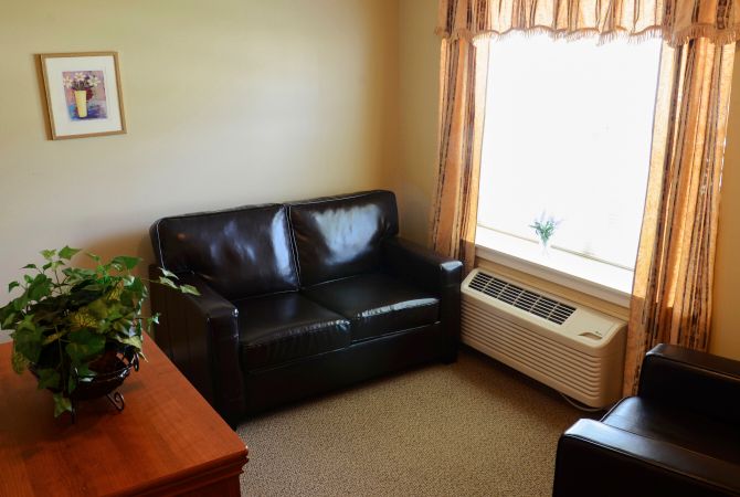 A cozy corner of a living room featuring a black leather sofa, adjacent to a window with curtains and an air conditioning unit.