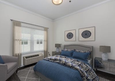 A modern bedroom with a bed covered in blue and patterned bedding, featuring matching pillows. Two pieces of artwork hang above the headboard. A lamp is situated on the bedside table near a window with blinds.