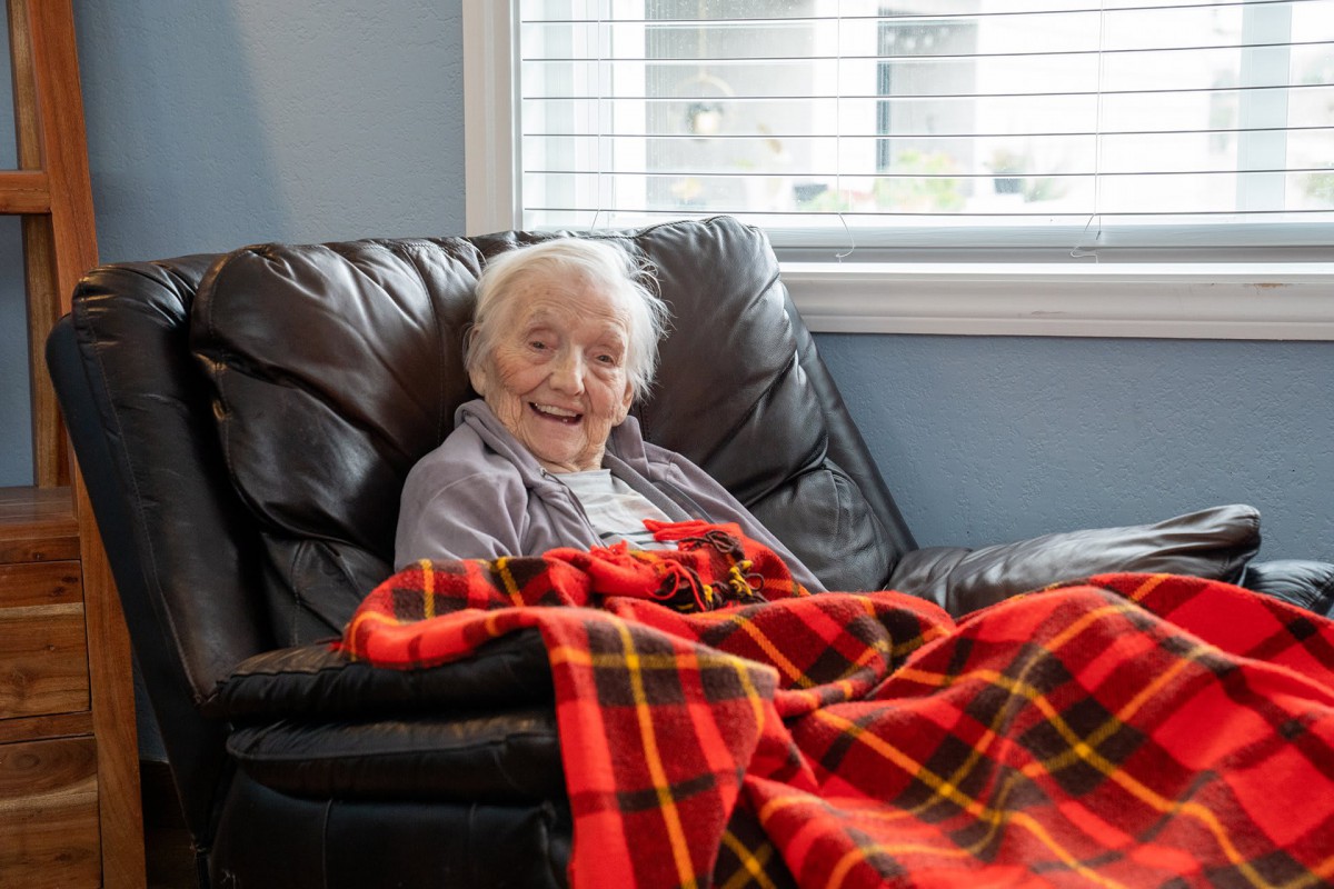 An elderly person smiling while sitting in a chair, covered with a warm plaid blanket near a window.