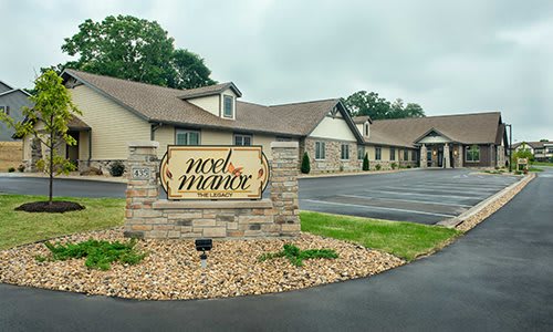 A view of a building with a sign reading 'Noel Manor' surrounded by greenery and a parking area.