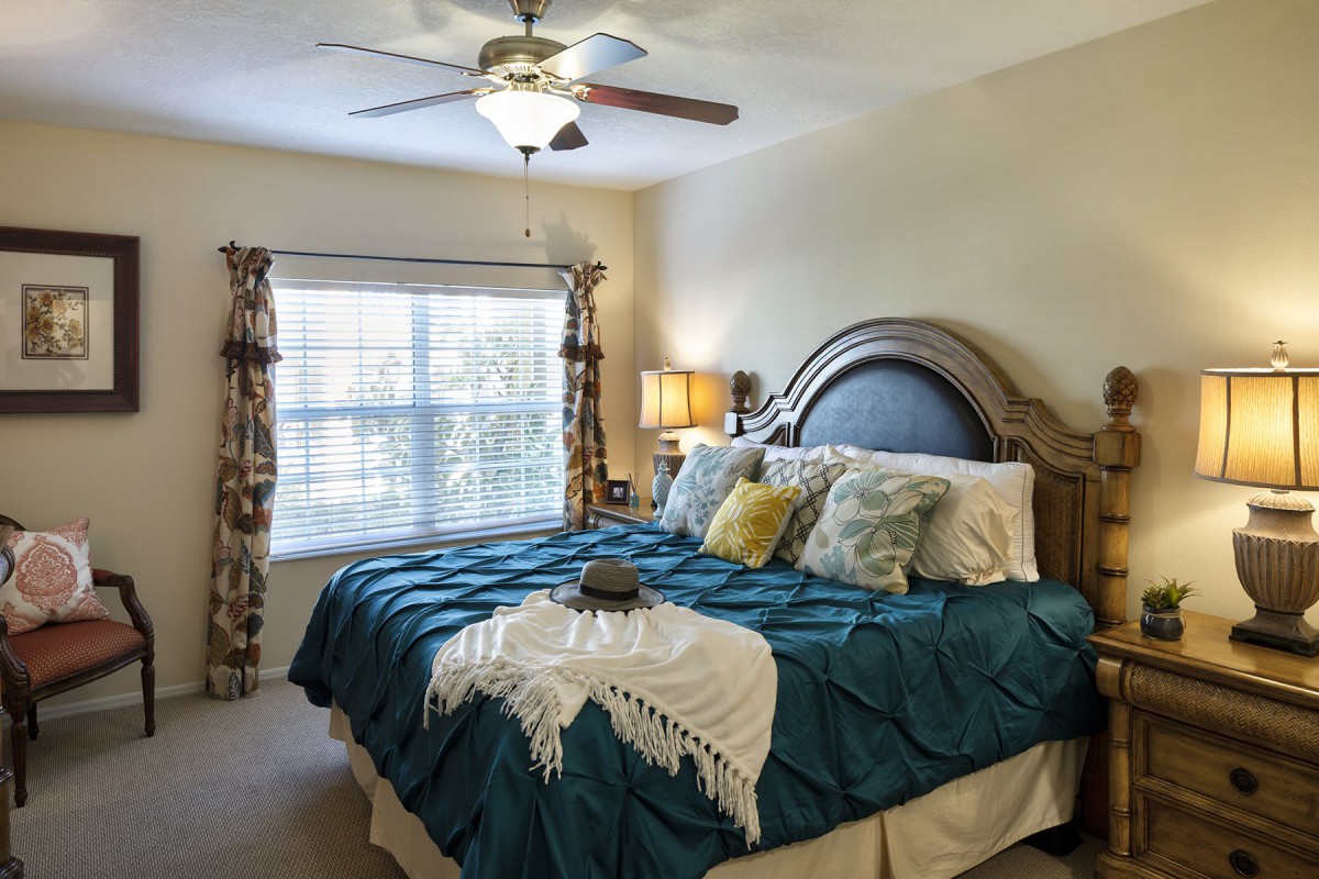 A cozy bedroom with a large bed adorned with decorative pillows and a throw blanket, featuring a window with patterned curtains and a ceiling fan.
