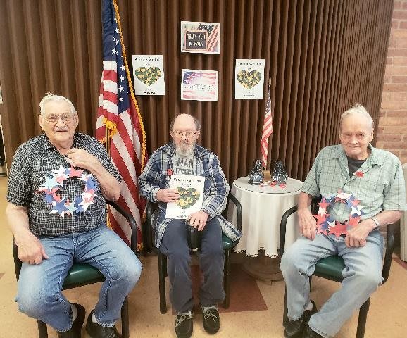 Three senior men are seated indoors, each wearing patriotic-themed attire with American flags in the background.