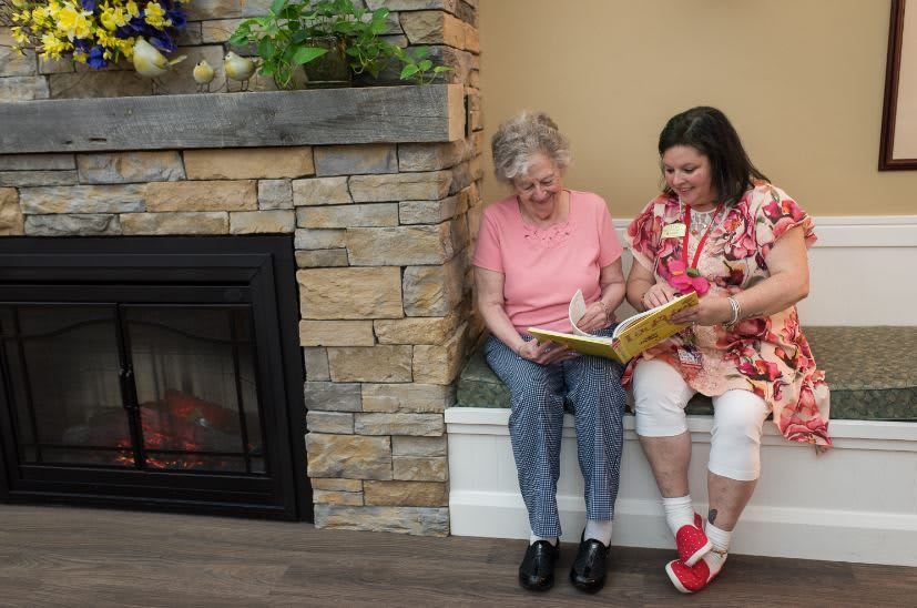An elderly woman and a younger woman sit together by the fireplace, enjoying a book.