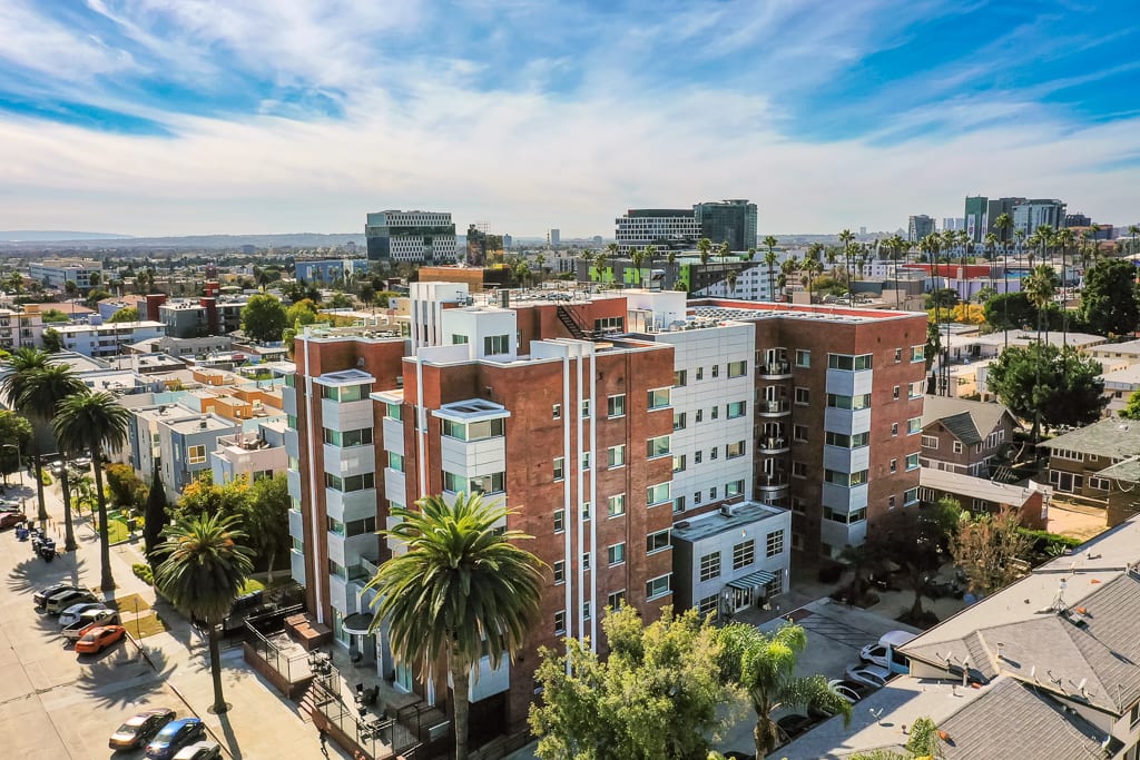 Aerial view of a modern apartment building in an urban setting with palm trees and city skyline in the background.