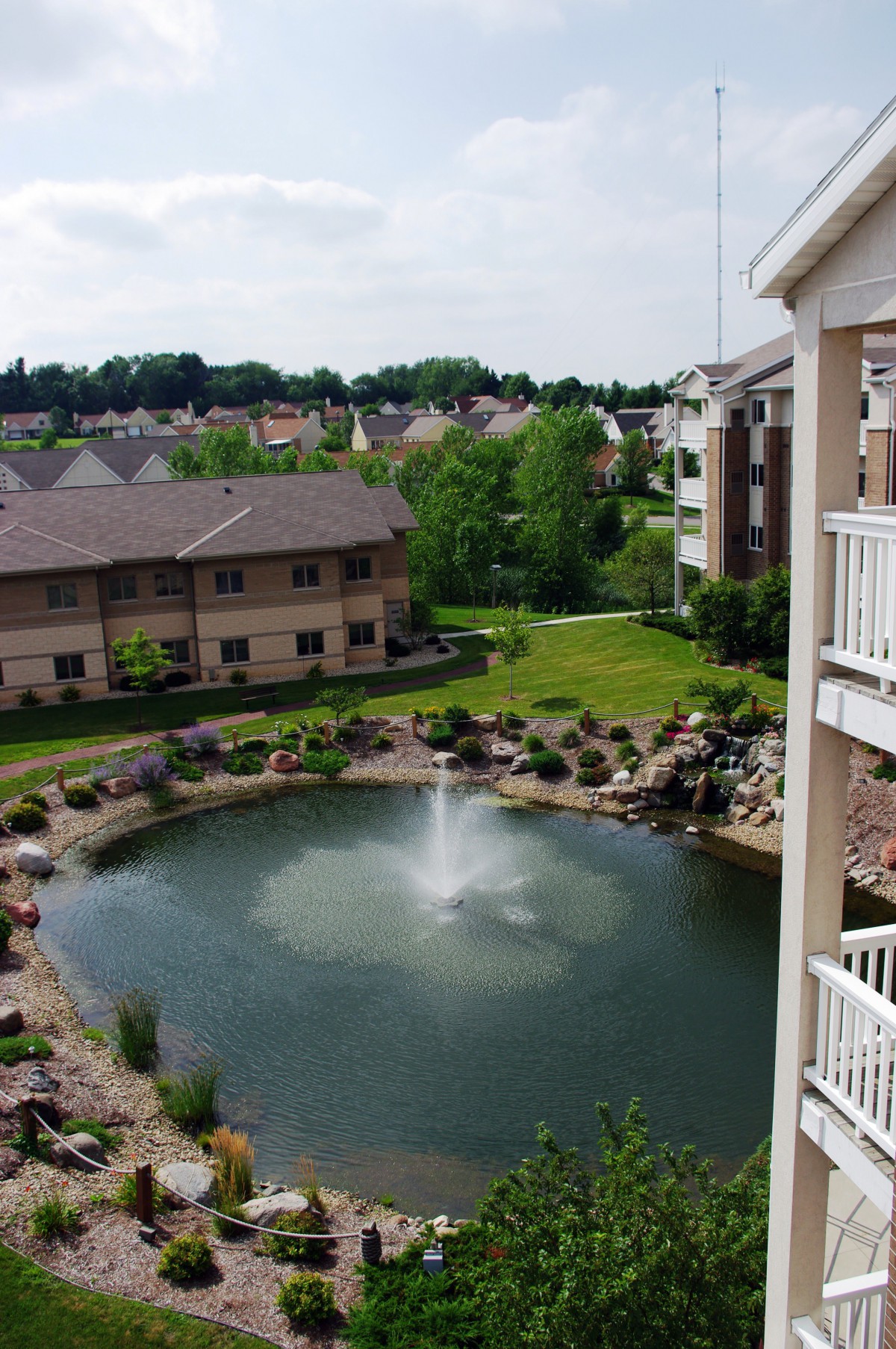 A scenic view of an apartment complex featuring a central pond with a fountain surrounded by greenery.