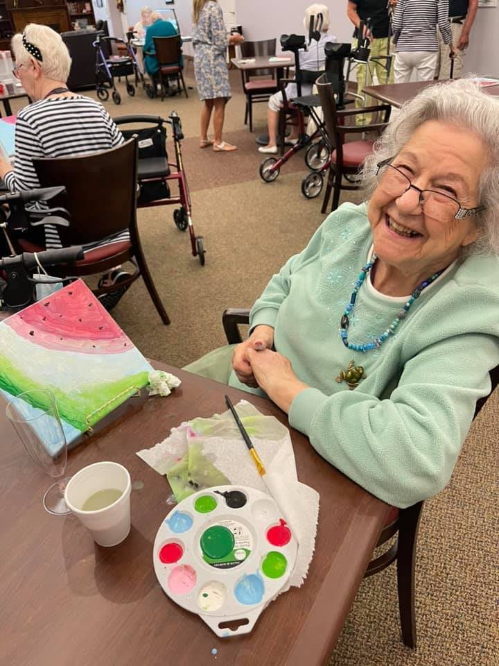 An elderly woman happily painting at a retirement home.