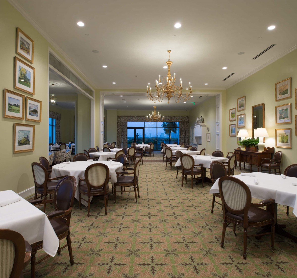 A well-lit, elegant dining room with neatly arranged tables and chairs, adorned with chandeliers and artwork on the walls.