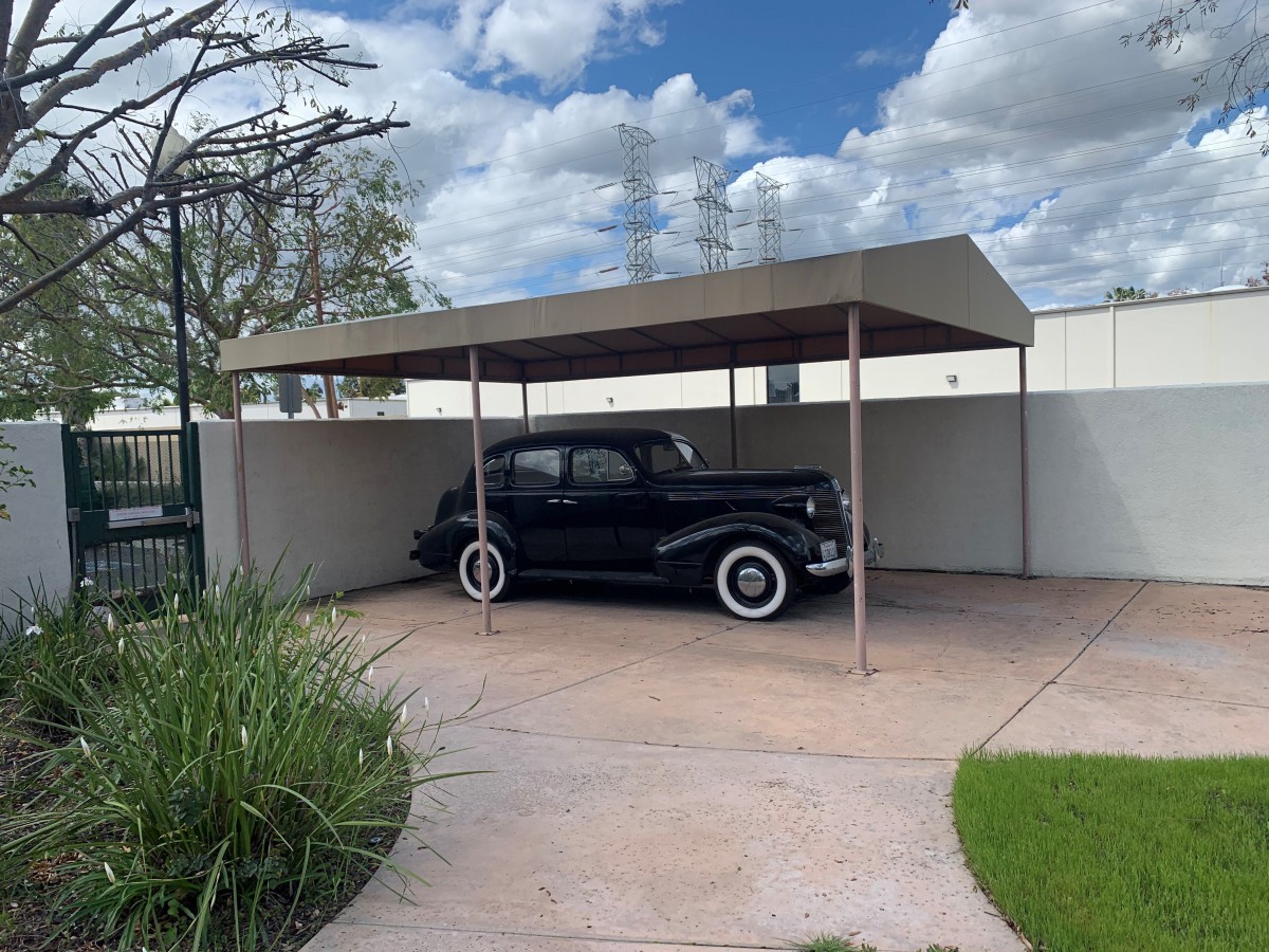 A classic vintage car parked under a modern carport on a partly cloudy day.