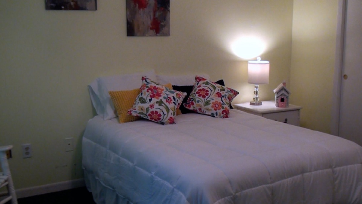 A cozy bedroom with a neatly made bed adorned with floral patterned pillows and a softly lit lamp on the nightstand.