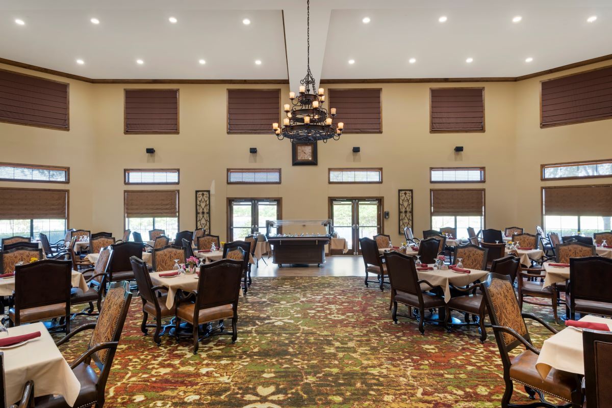 An elegant dining room with neatly arranged tables and chairs, featuring a central chandelier and large windows.