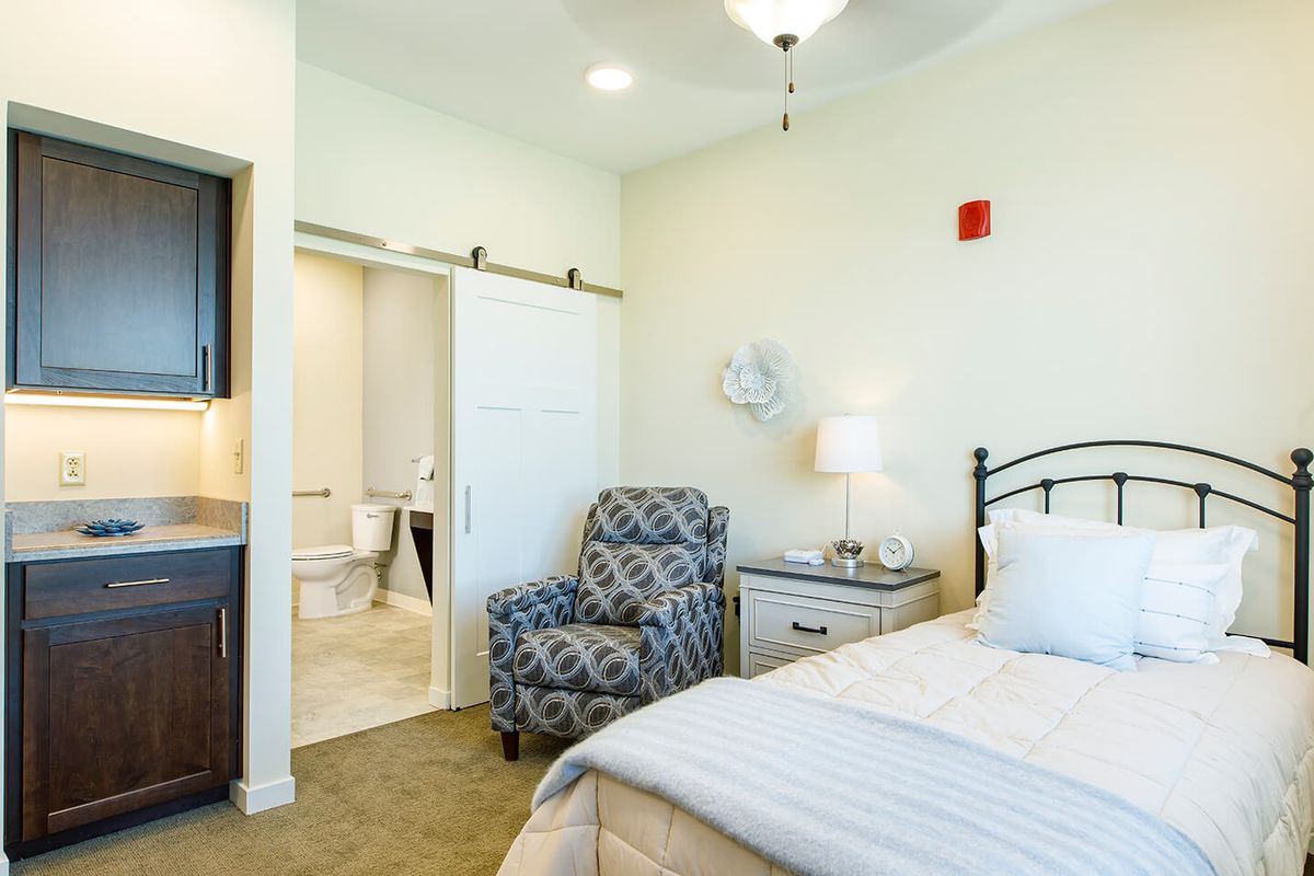A cozy bedroom with white walls and a comfortable bed, featuring a patterned armchair, nightstand, and lamp.