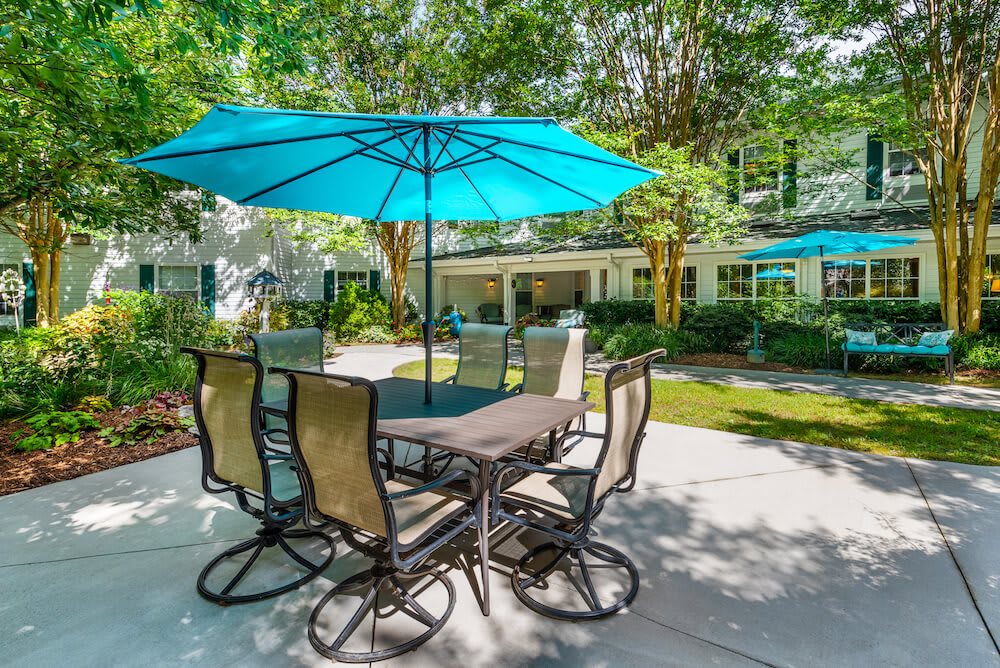 A sunny outdoor patio with a table, chairs, and a blue umbrella surrounded by greenery.