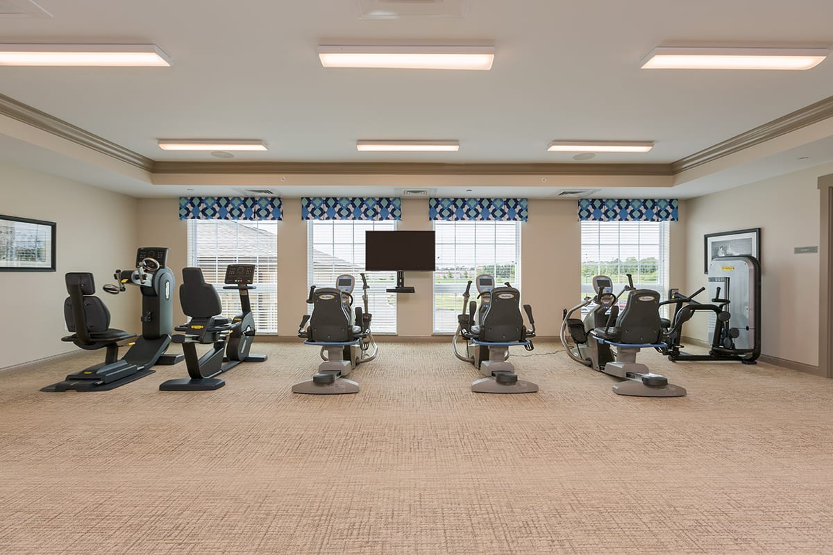 A modern gym with stationary bikes and treadmills, featuring large windows and a central TV.