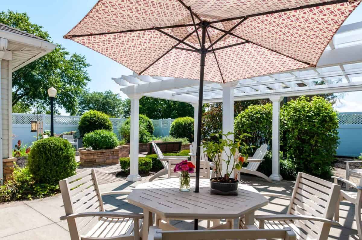 A sunny patio with a wooden table and chairs under a large umbrella, surrounded by lush green landscaping.