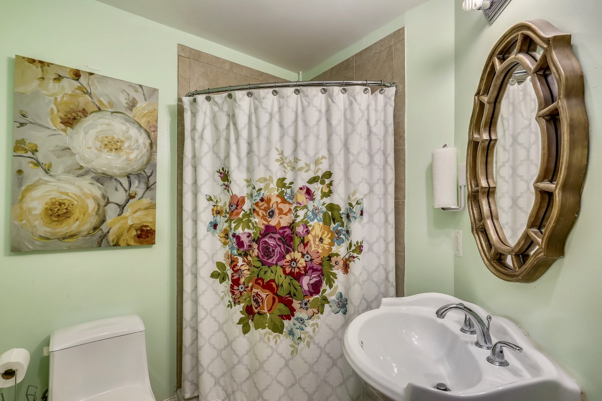 A cozy bathroom with a floral shower curtain, pastel green walls, and decorative artwork.