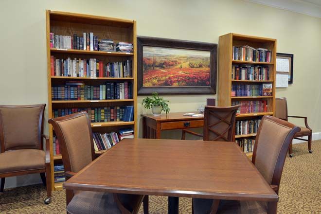 A quiet reading area in a library with bookshelves, a wooden table and chairs, and a landscape painting on the wall.