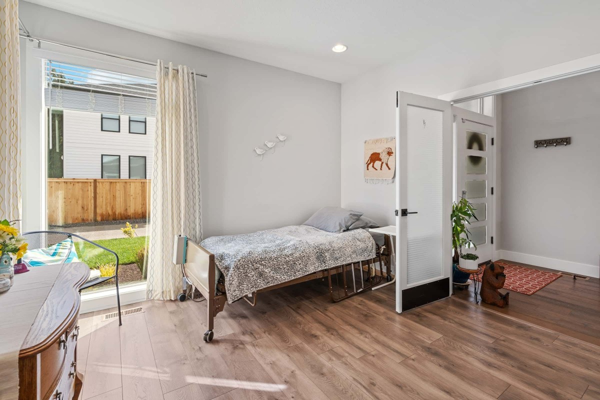 A minimalist bedroom with natural light, featuring a single bed, wood flooring, and simple decor.