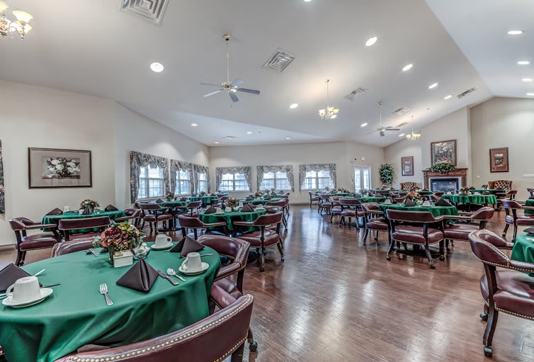 A spacious dining room with neatly arranged tables covered in green tablecloths, surrounded by comfortable chairs and well-lit by ceiling lights.