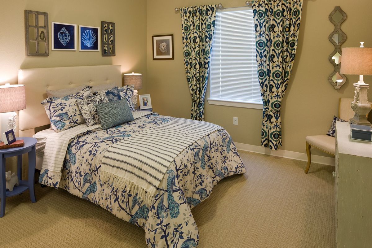 A cozy bedroom with floral patterned bedding and curtains, featuring a bedside lamp and framed artwork on the walls.