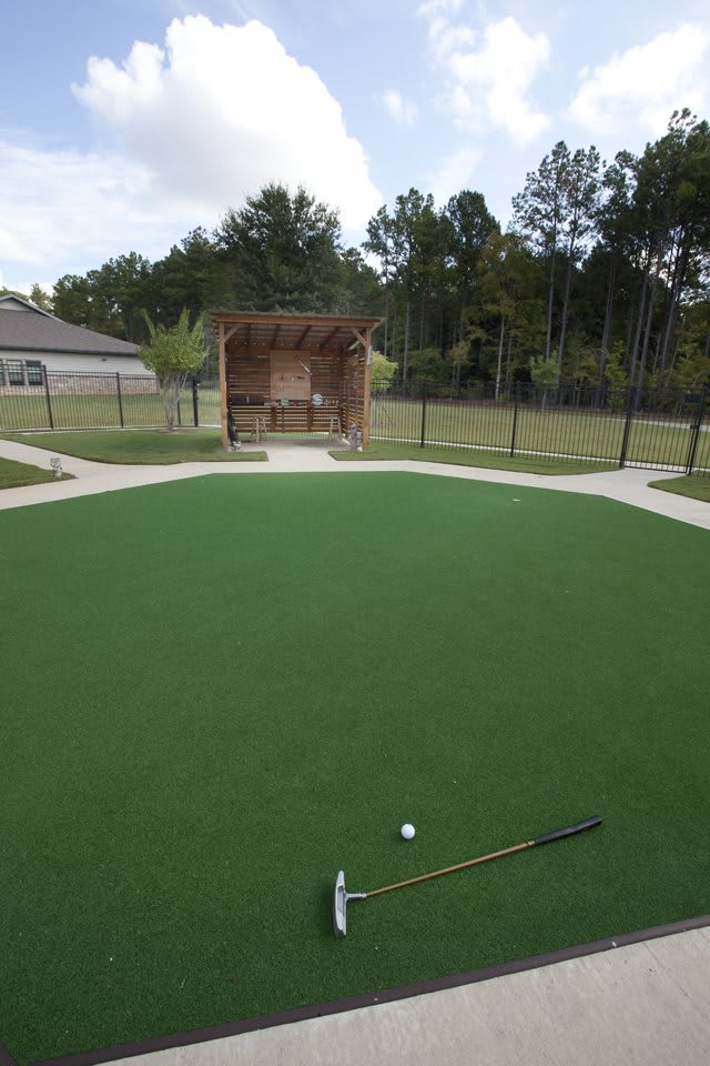 A well-maintained outdoor putting green with golf clubs and a ball, surrounded by fencing and a small shelter in the background.