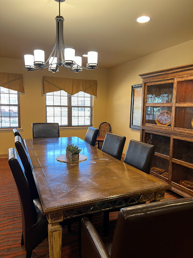 A spacious dining room featuring a large wooden table, black chairs, and a stylish chandelier, illuminated by natural light from wide windows.