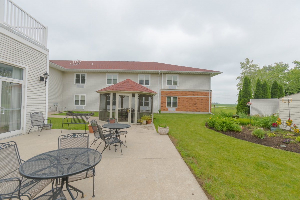 A two-story building with a patio featuring outdoor furniture, surrounded by a garden under an overcast sky.