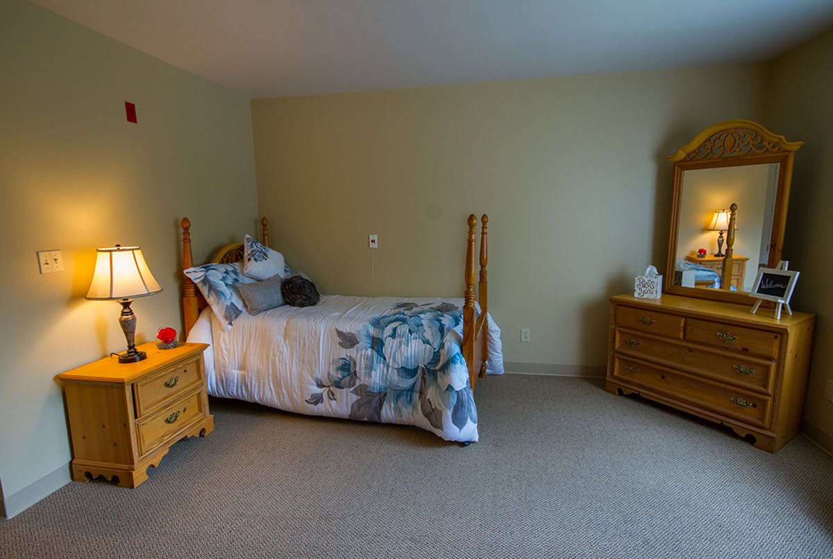 A cozy bedroom featuring a single bed with wooden posts, adorned with decorative pillows and a bedside lamp, alongside a dresser with a mirror.