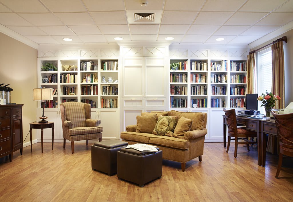 A cozy library reading area with bookshelves, a sofa, armchair, and wooden flooring.
