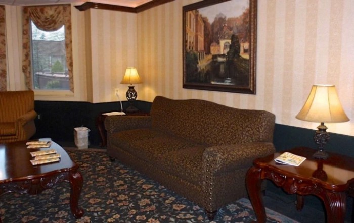 A cozy living room with a brown sofa, side tables, and a lamp, decorated with a large painting and patterned carpet.