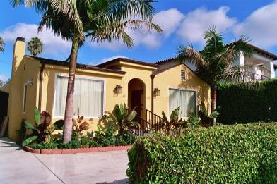 A tropical-style yellow house surrounded by palm trees and lush landscaping on a sunny day.