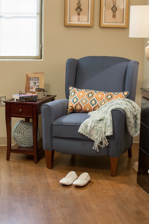 A cozy living room corner with a blue armchair and a decorative pillow, complemented by a side table and slippers on the floor.
