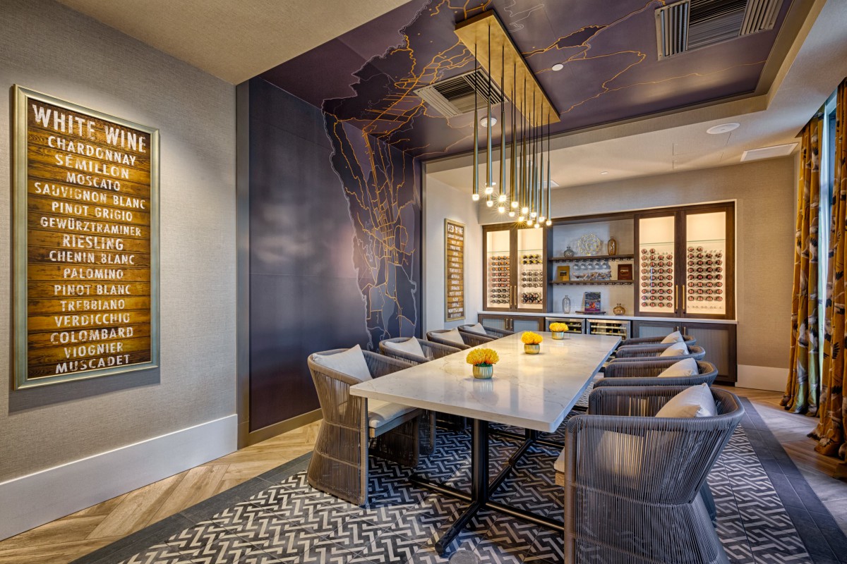 A modern dining room with elegant decor, featuring a long table, cushioned chairs, a stylish chandelier, and wine storage along the walls.