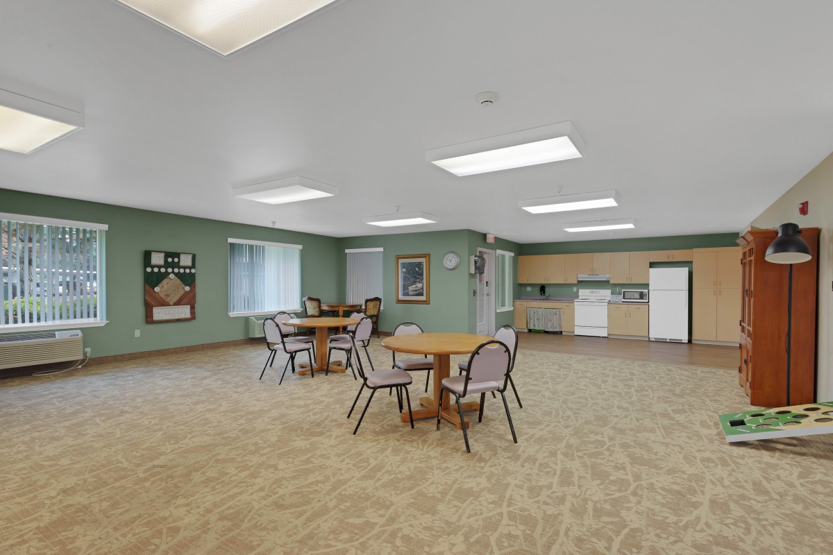 An empty room with tables and chairs, featuring a small kitchen area and windows.
